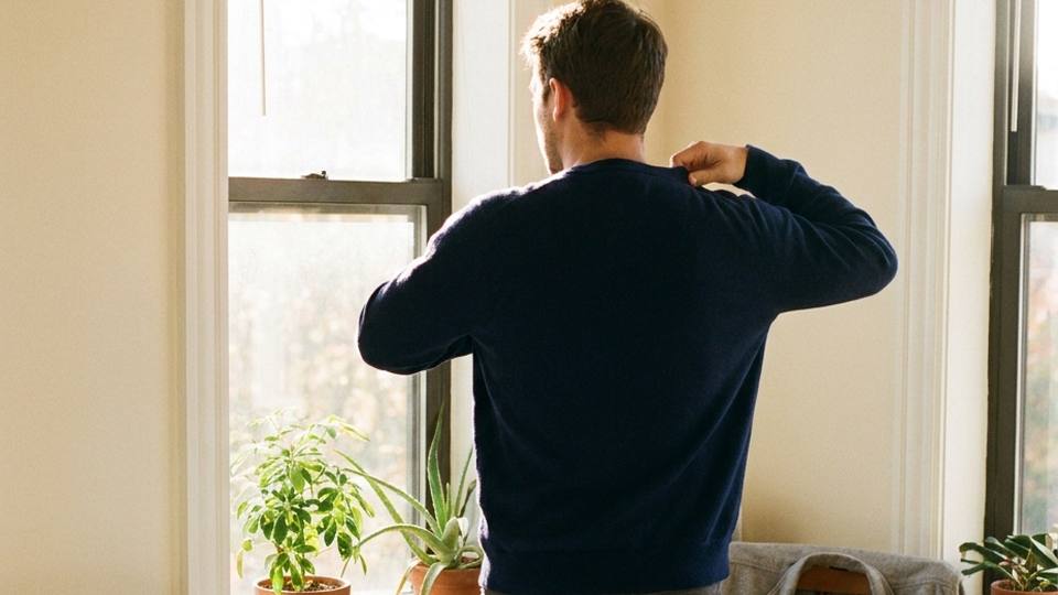 A man pulls on a navy sweater by a sunny bedroom window